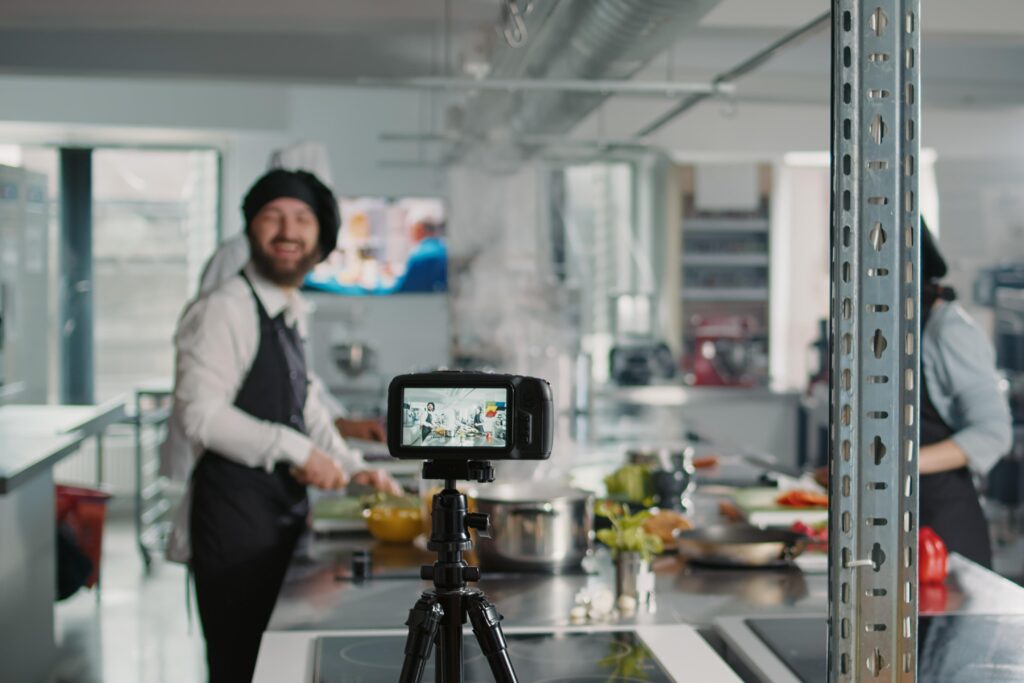 In a professional kitchen, a digital camera on a tripod is positioned to record or stream chefs at work, with the live feed visible on its small screen. In the background, a smiling chef in an apron and hat prepares food.