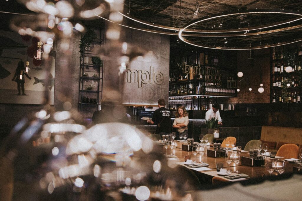 A sophisticated, dimly lit restaurant interior features a long wooden dining table set with elegant glassware under glowing circular chandeliers. The shot uses a shallow depth of field, with blurred foreground elements framing a view of staff and patrons near a modern bar.