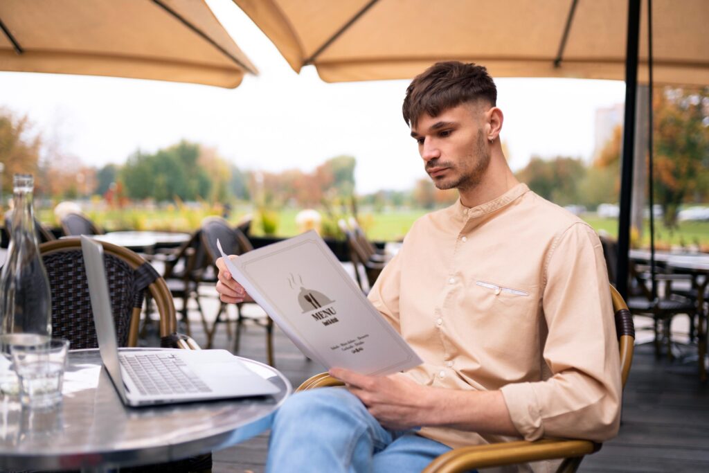A man in a beige shirt sits at an outdoor cafe table, carefully reviewing a handheld menu. An open laptop, a glass of water, and a bottle are positioned on the table beside him.