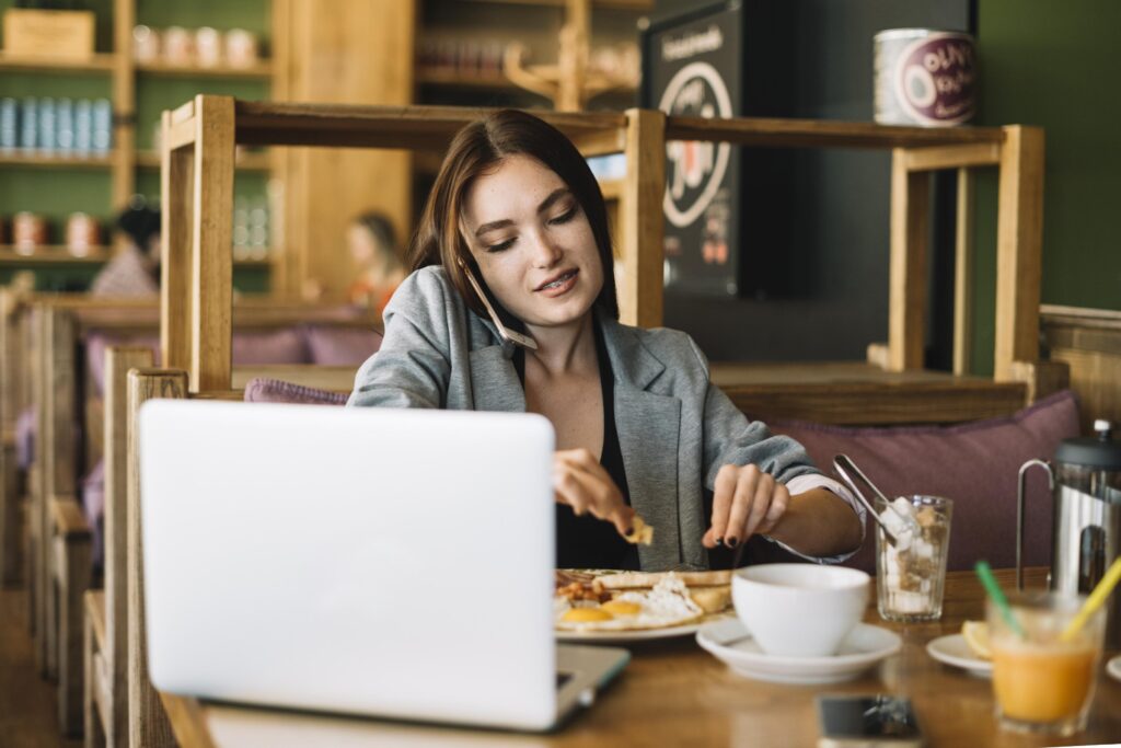 A woman in a grey blazer talks on her phone while having breakfast and working on a laptop at a cafe. The scene captures a busy morning atmosphere with a plate of eggs, toast, and coffee spread across the wooden table.