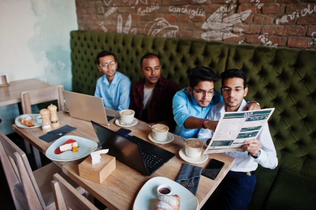 Four men are seated at a long wooden table in a cafe, working on laptops and reviewing a menu together. The table is spread with coffee, pastries, and desserts against a backdrop of a green tufted booth and a brick wall.