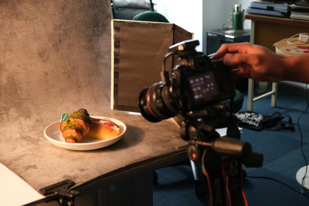 A camera on a tripod is positioned to capture a professional shot of a croissant and pastry on a white plate. The setup includes a textured gray backdrop and a light reflector to enhance the food's appearance.