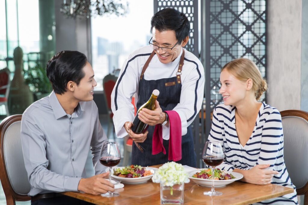 A smiling waiter in a white chef's coat and denim apron presents a bottle of wine to a young couple seated at a restaurant table. The couple looks up at him with interest while enjoying salads.