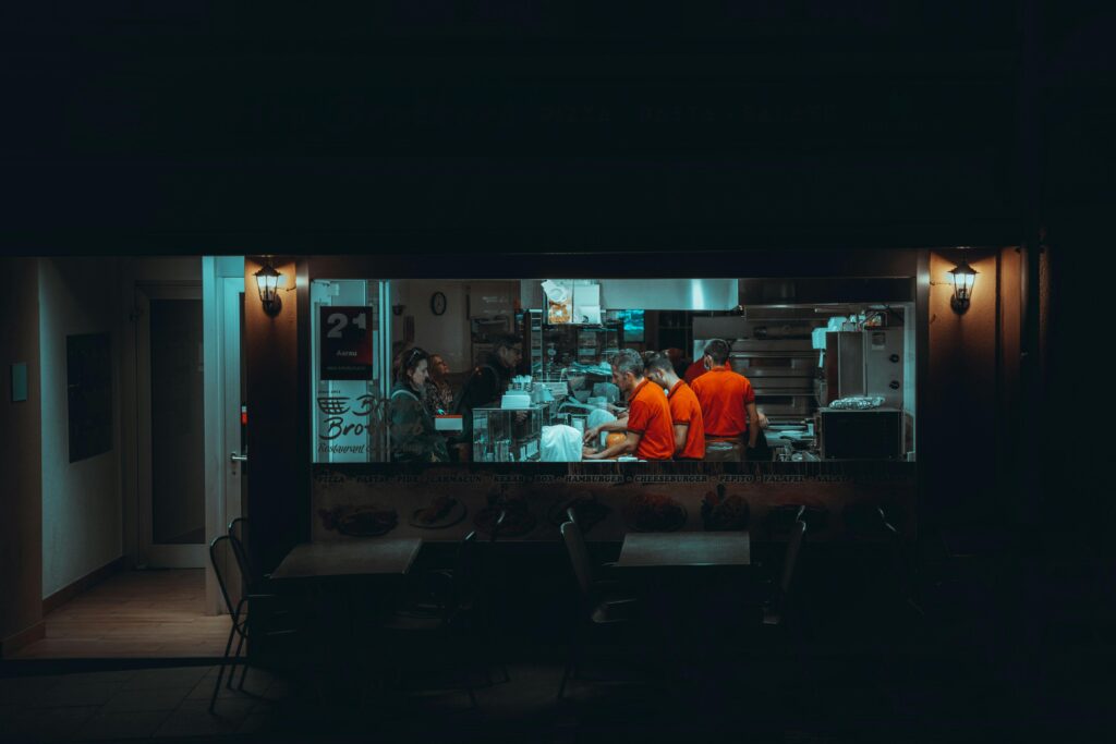 A glowing restaurant window illuminates several workers in orange shirts as they prepare food in a dark kitchen. Outside, empty tables sit in the shadows of the night.