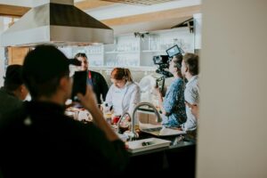 A professional film crew is recording a chef preparing food in a modern kitchen. Several people are observing and filming the process from behind a counter.