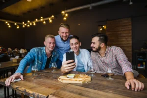 Four men are gathered around a wooden table in a dimly lit restaurant, smiling and laughing as they look at a smartphone held by one of them. The table is set with short-stemmed glasses containing an amber beverage and a wooden platter with a snack and dip.