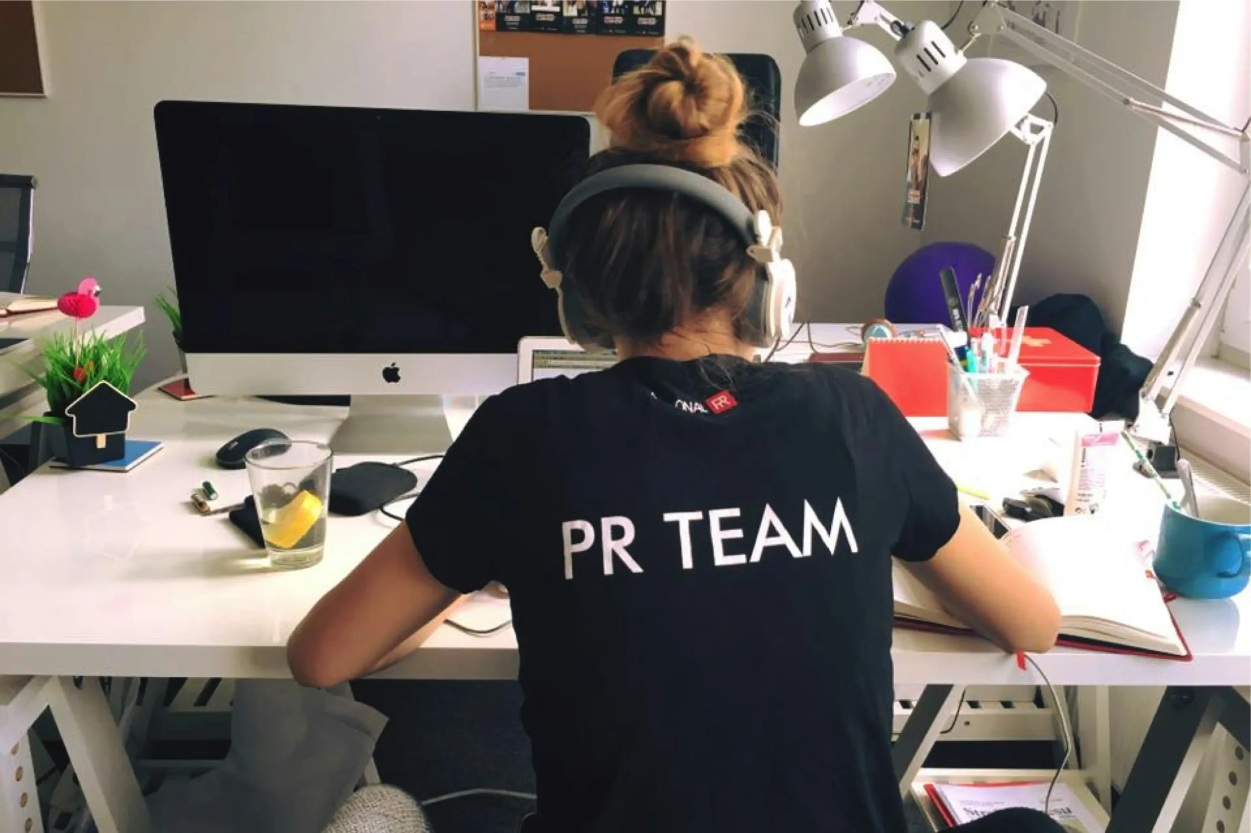A woman with her hair in a bun and wearing headphones sits at a white desk facing away from the camera, wearing a black T-shirt that says "PR TEAM". Her workspace is filled with a computer monitor, an open notebook, desk lamps, and various office supplies.