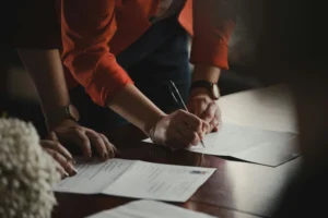 A person in an orange blazer is shown leaning over a wooden table to sign a document with a black pen. Another individual stands nearby with their hands resting on the table, observing the formal signing process.