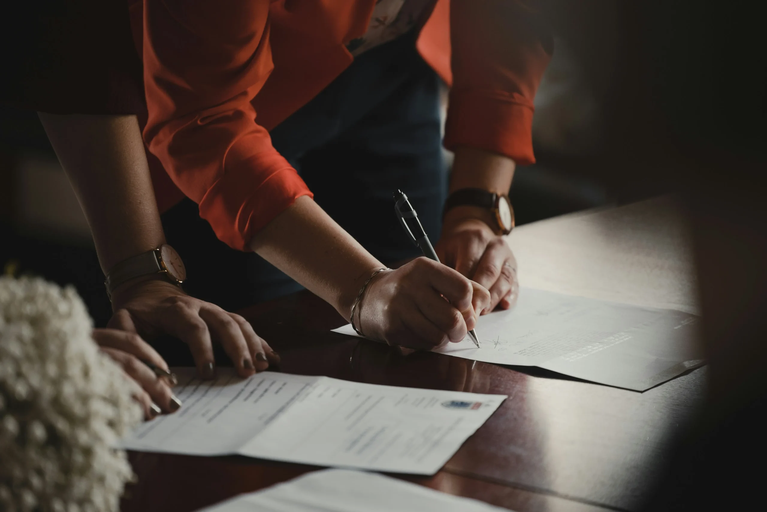 A person in an orange blazer is shown leaning over a wooden table to sign a document with a black pen. Another individual stands nearby with their hands resting on the table, observing the formal signing process.