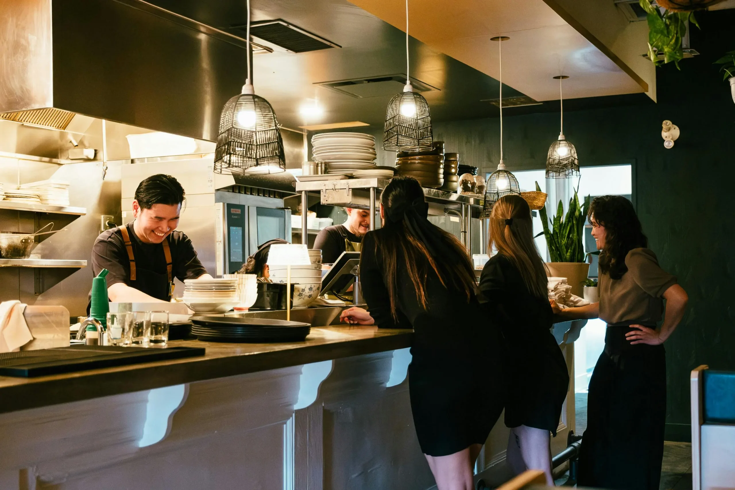 A chef smiles while working in an open kitchen as several patrons lean against the counter and engage in conversation. The restaurant features a modern, moody atmosphere highlighted by woven pendant lights and dark green walls.