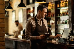 A smiling barista or bartender in a brown apron stands behind a wooden counter, holding a small tablet while interacting with a POS touchscreen system. In the warm, softly lit background, another worker is visible near shelves stocked with bottles.
