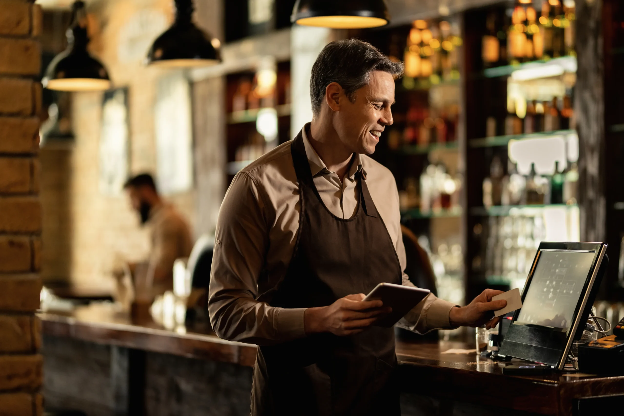 A smiling barista or bartender in a brown apron stands behind a wooden counter, holding a small tablet while interacting with a POS touchscreen system. In the warm, softly lit background, another worker is visible near shelves stocked with bottles.