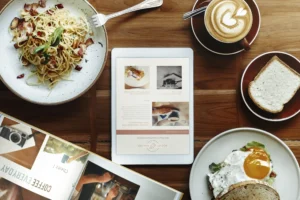 An overhead shot displays a wooden table arranged with a plate of pasta, a fried egg sandwich, a slice of bread, a cup of latte art coffee, and an open magazine. In the center, a tablet screen shows a webpage titled "ROCKET ROASTER" alongside articles about vintage gadgets and photography.