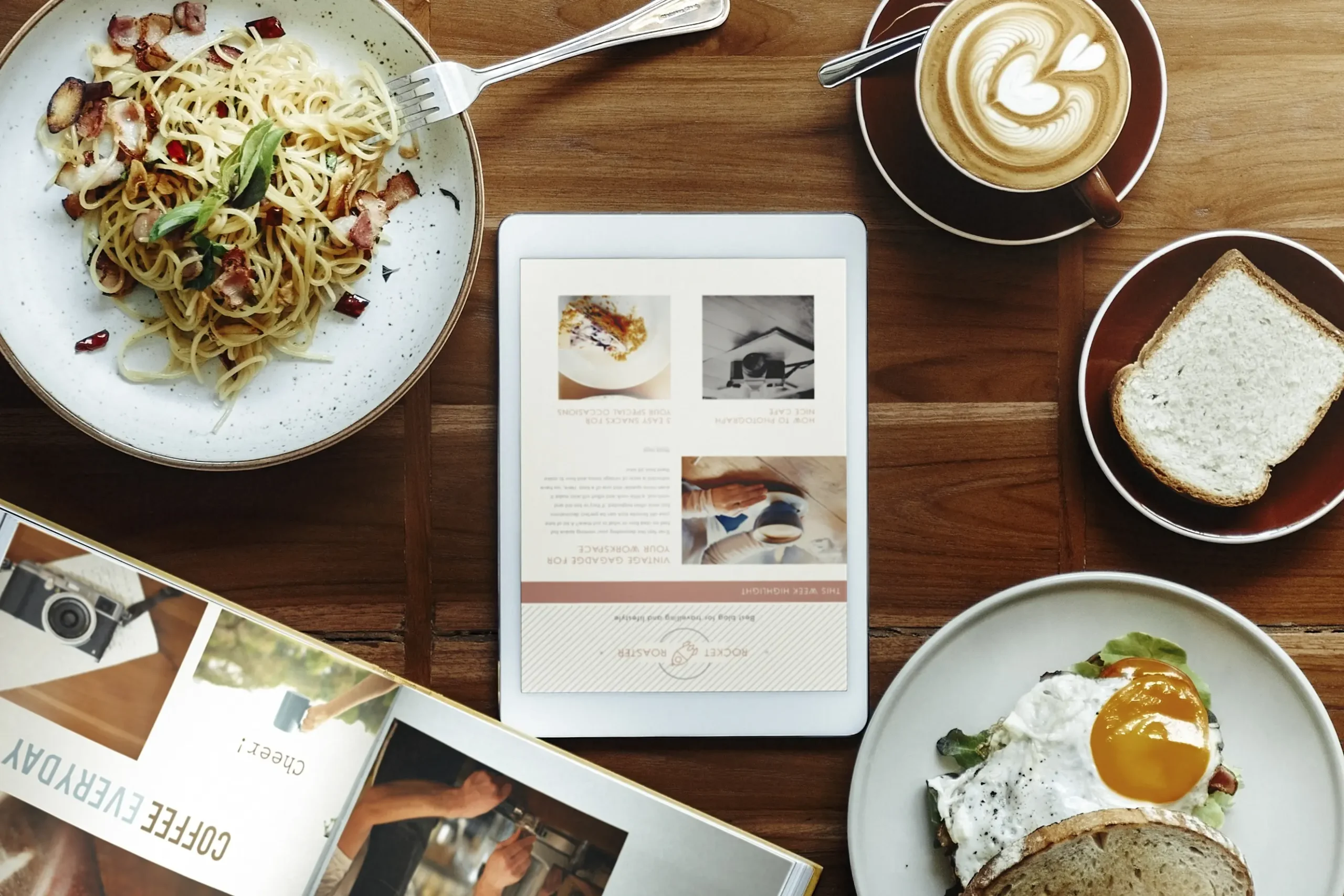 An overhead shot displays a wooden table arranged with a plate of pasta, a fried egg sandwich, a slice of bread, a cup of latte art coffee, and an open magazine. In the center, a tablet screen shows a webpage titled "ROCKET ROASTER" alongside articles about vintage gadgets and photography.