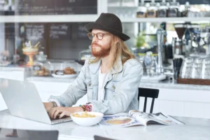 A man with long hair, a beard, a hat, and glasses is sitting at a cafe table while working on a laptop. Beside him on the marble surface are a bowl of french fries and an open magazine.