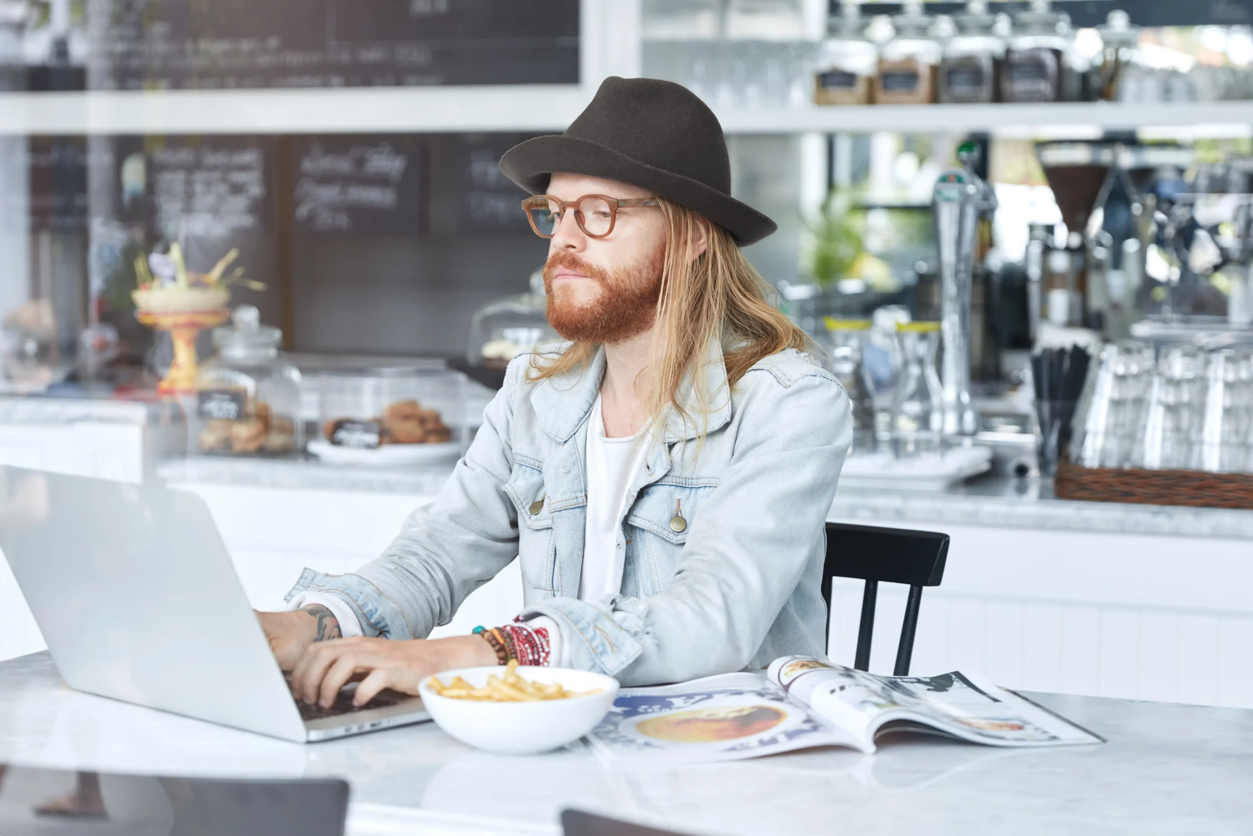 A man with long hair, a beard, a hat, and glasses is sitting at a cafe table while working on a laptop. Beside him on the marble surface are a bowl of french fries and an open magazine.