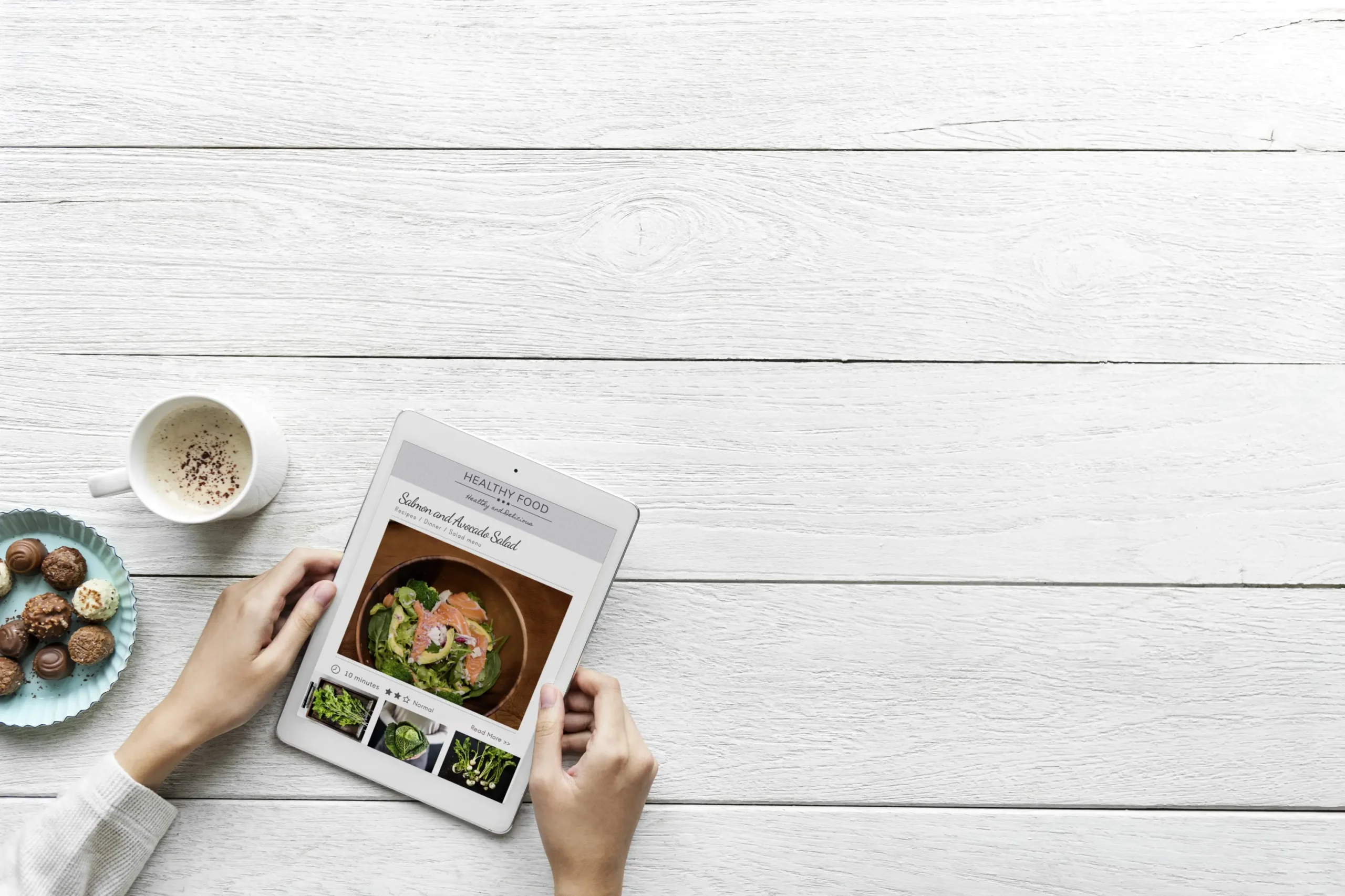 Hands hold a tablet displaying a salmon and avocado salad recipe on a white wooden surface. A cup of coffee and a small plate of chocolate truffles sit alongside the device to complete the flat-lay scene.