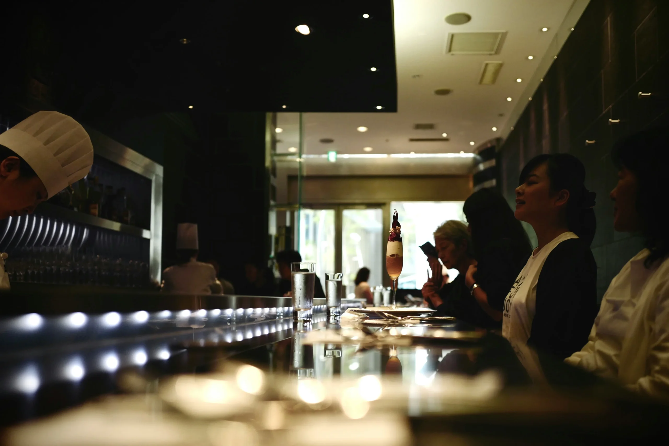 A chef in a white toque works diligently behind a sleek, modern restaurant counter lined with soft, glowing LED lights. On the opposite side, several patrons sit in the dimly lit space, watching the kitchen or enjoying their drinks in an intimate atmosphere.
