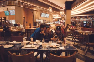 A family huddles together at a marble table in a brightly lit, modern food court to look at a smartphone. The image captures a warm, candid moment amidst the bustling background of other diners and a nearby casino area.