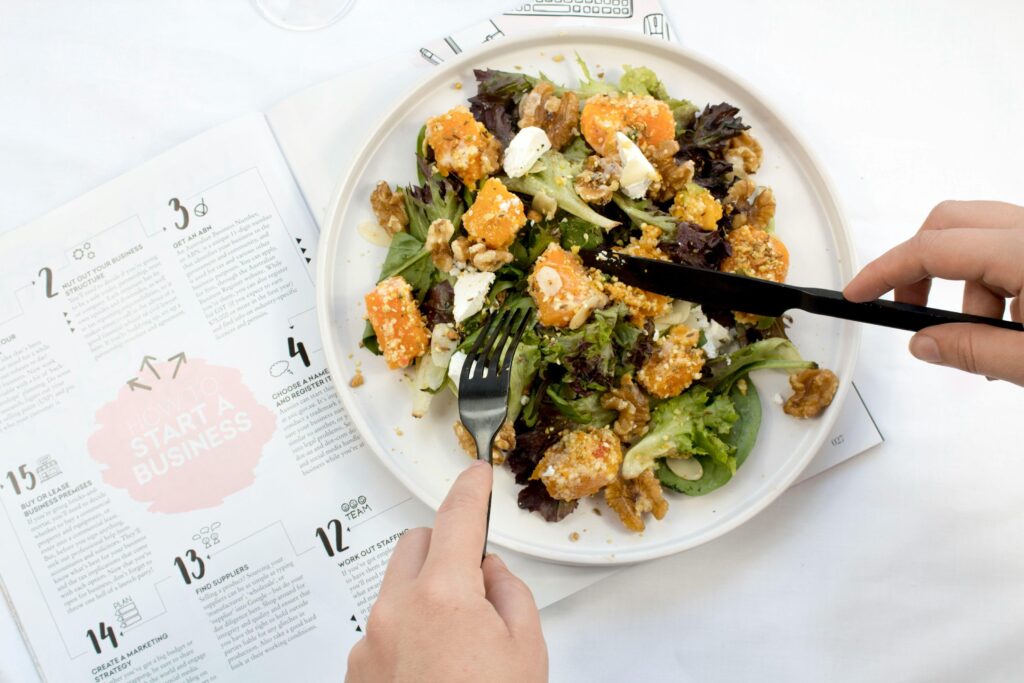 A vibrant salad featuring roasted pumpkin, walnuts, and greens is served on a white plate. Hands use a black fork and knife to eat the meal atop an open magazine about starting a business.