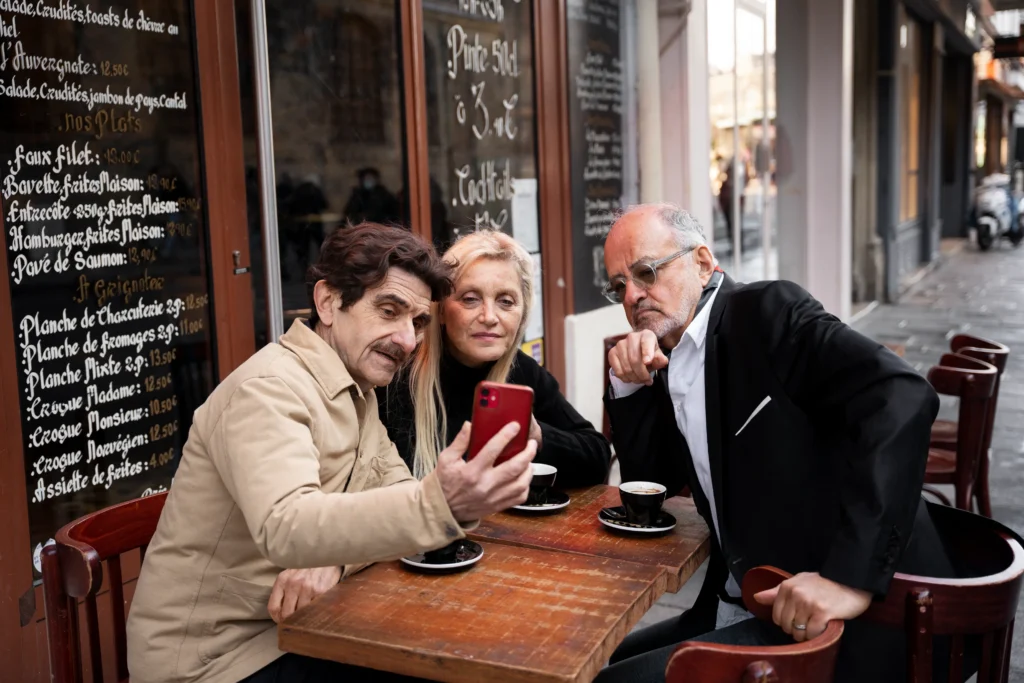 Three older friends are sitting at an outdoor cafe table in front of a French menu board, looking at a red smartphone held by one of the men. The man holding the phone wears a tan jacket, the woman in the middle looks on intently, and the man on the right in a black blazer leans in while squinting at the screen.