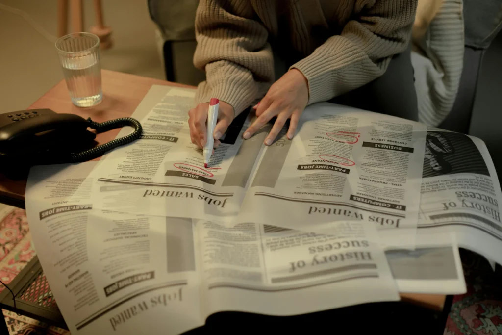 A person is shown from the chest down, wearing a beige sweater and using a pink highlighter to circle listings in the "Jobs Wanted" section of a newspaper. The newspaper pages are spread out on a wooden coffee table next to a landline telephone and a glass of water.