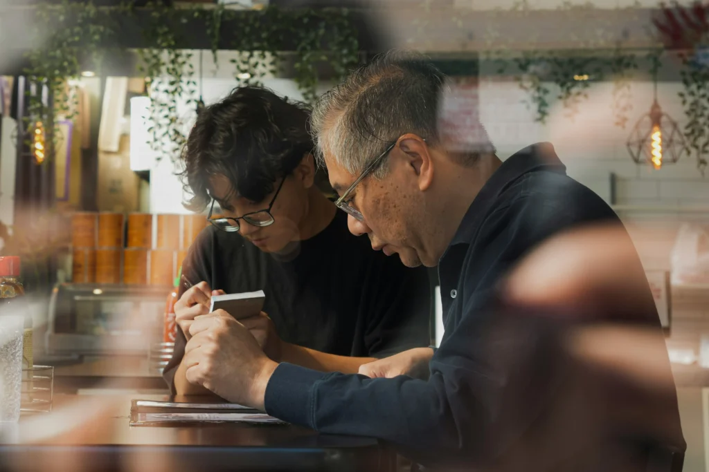 Two men are seated at a counter, intently looking down at a menu and a notepad as if preparing to place an order. The scene is captured through a glass window with soft reflections, creating an intimate atmosphere within a cozy, dimly lit eatery.