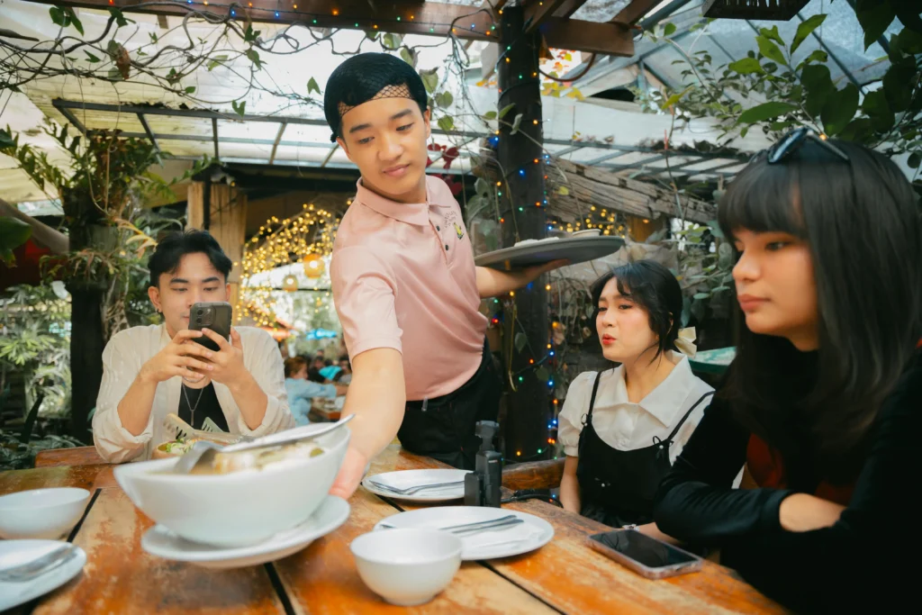 A waiter in a pink polo shirt and hairnet serves a large bowl of steaming food to three young diners seated at an outdoor wooden table. The lush, garden-like setting is decorated with hanging vines and warm fairy lights, creating a cozy and rustic atmosphere.