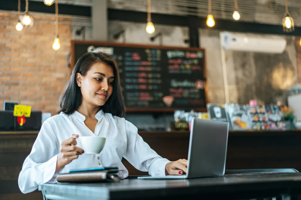 A woman sits at a table in a cozy coffee shop, holding a white mug while working on a laptop. The background features warm industrial decor, including exposed brick walls, hanging light bulbs, and a menu board.
