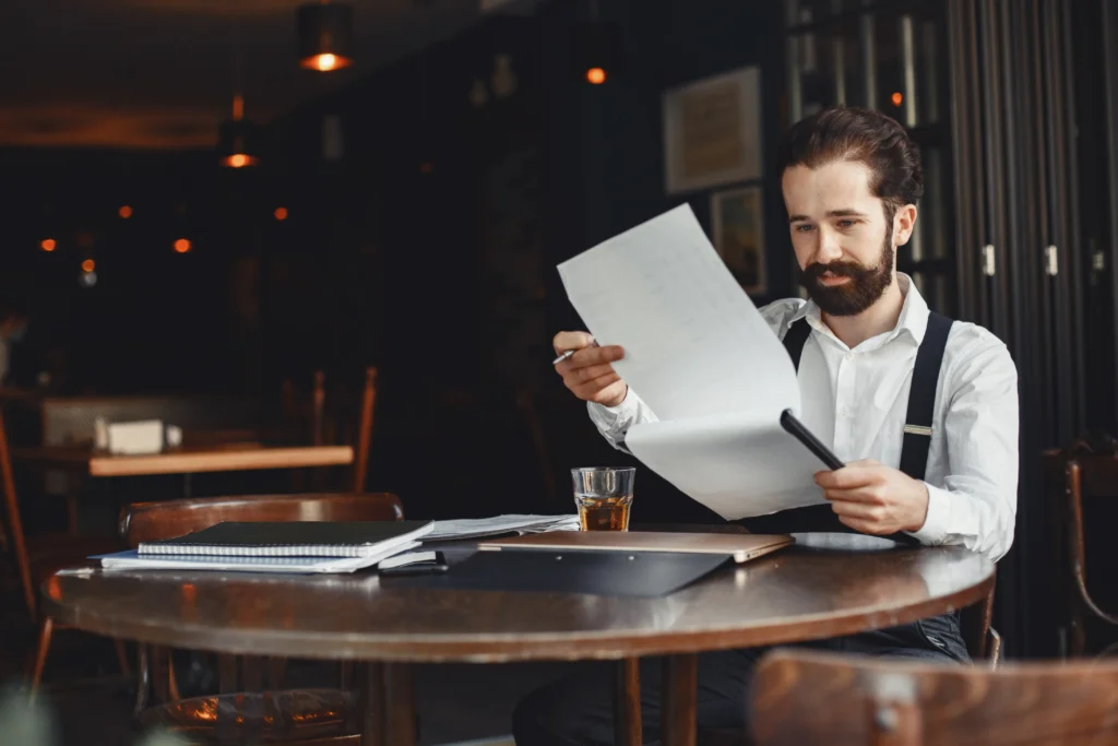 A bearded man wearing a white shirt and suspenders sits at a round wooden table reviewing several documents. A glass of amber liquid and various notebooks rest on the table before him in the dimly lit, moody setting.