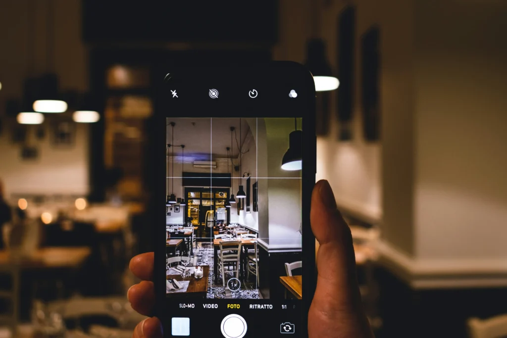 A person's hand holds a smartphone, capturing a photo of a dimly lit restaurant interior with tables, chairs, and hanging lights. The phone screen displays the camera interface with grid lines framing the warm, cozy dining space.