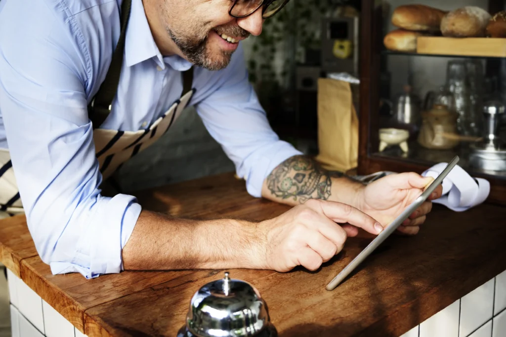 A smiling, tattooed baker wearing a striped apron leans over a wooden counter while using a digital tablet. In the background, a display case is filled with fresh bread loaves and various coffee shop equipment.
