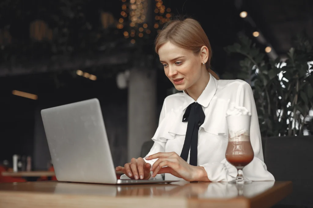 A woman in a white ruffled blouse with a black bow sits at a wooden table in a dimly lit cafe, focused on her laptop. Beside her sits a tall glass of coffee topped with whipped cream, while warm bokeh lights glow in the background.