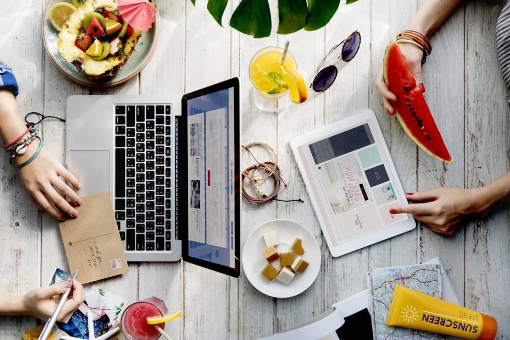 Several people are gathered around a white wooden table filled with travel essentials, including a laptop, tablet, sunscreen, and a map. The scene is accented by tropical touches like fresh fruit, colorful drinks, and a small pineapple bowl, suggesting they are planning or enjoying a vacation.