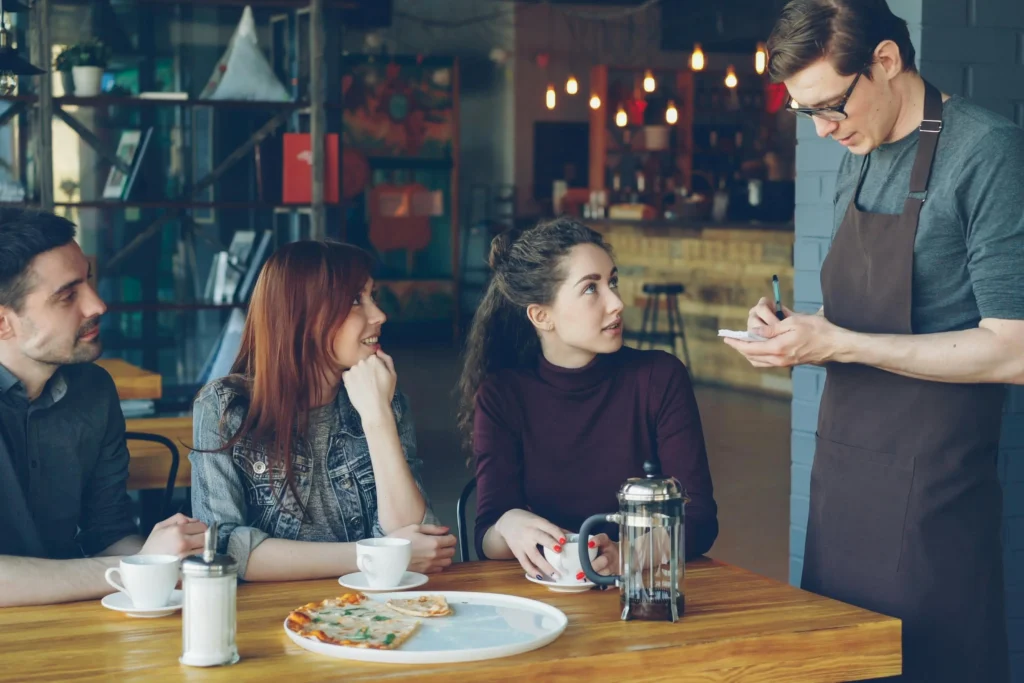 A waiter wearing an apron and glasses stands at a restaurant table, writing on a notepad as he takes an order from a group of three friends. A man and two women sit at the wooden table, which holds coffee cups, a French press, a sugar shaker, and a partially eaten pizza on a plate.