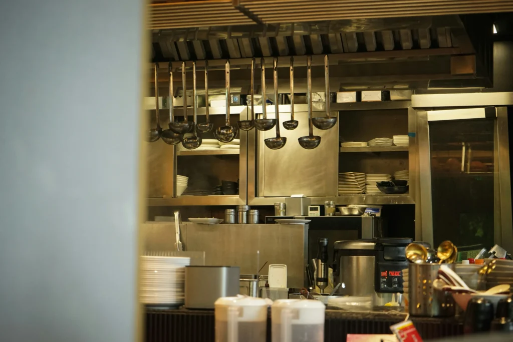 The image shows a professional commercial kitchen featuring stainless steel surfaces and various cooking equipment. Numerous metal ladles hang from an overhead rack above a counter stacked with white plates and kitchen appliances.