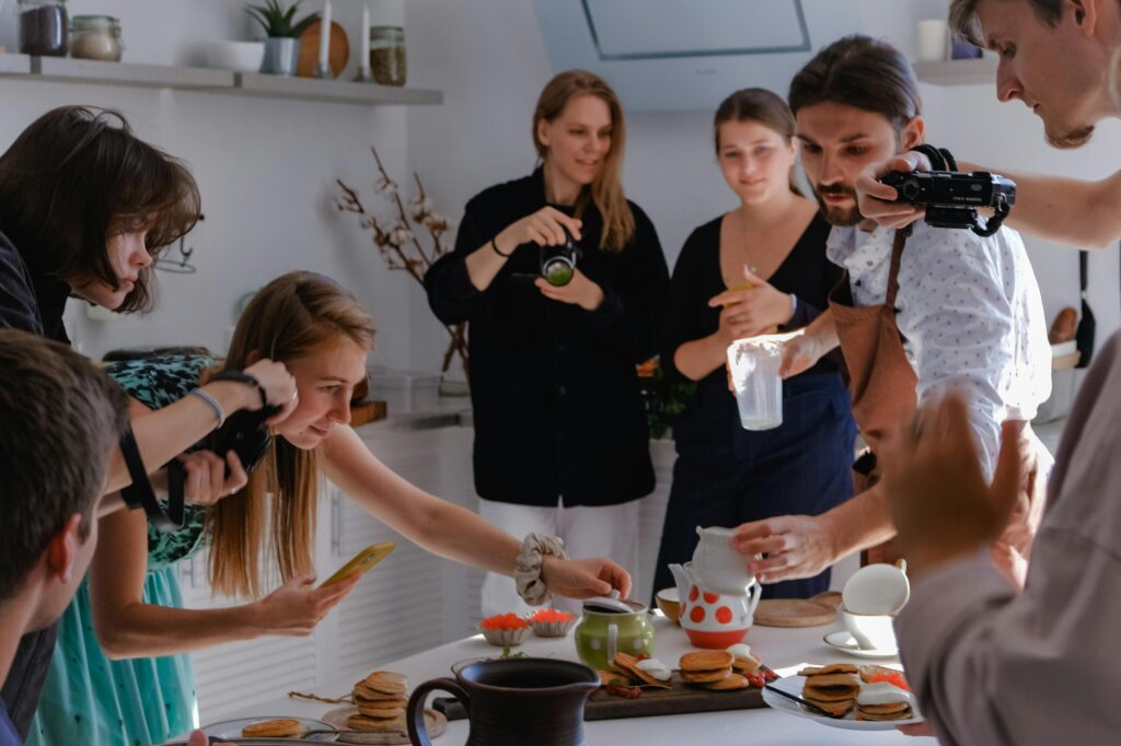 A group of people gathers in a bright kitchen to photograph a meal being prepared. Everyone is focused on capturing the food and tea service with their cameras and phones.