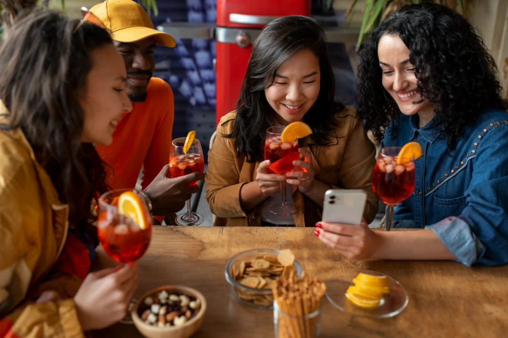 A diverse group of four friends sits around a wooden table, smiling and looking at a smartphone together while holding orange-garnished spritz cocktails. The table in front of them is filled with various bar snacks, including pretzels, crackers, and nuts.