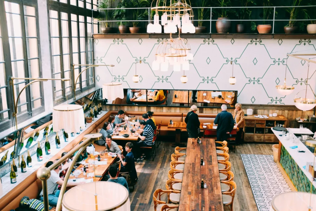 An aerial, wide-angle shot shows patrons dining in a spacious, trendy restaurant with high ceilings, long wooden tables, and large windows. The interior features geometric wall tiles, numerous hanging light fixtures, and a balcony lined with potted green plants.