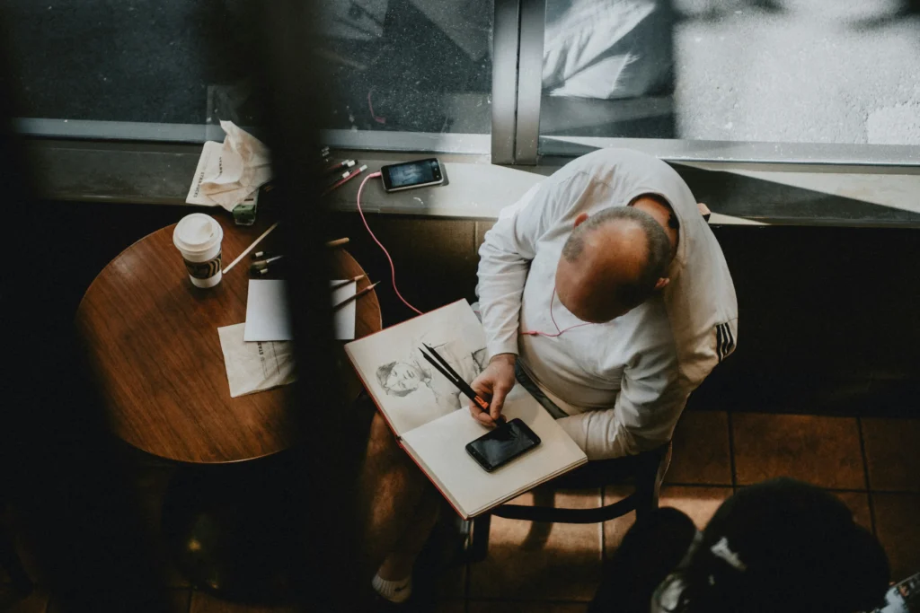 An artist sits at a small wooden table in a cafe, looking down as they sketch a portrait in a large sketchbook. The scene is captured from a high angle, showing pencils, a mobile phone, and a coffee cup scattered across their workspace.