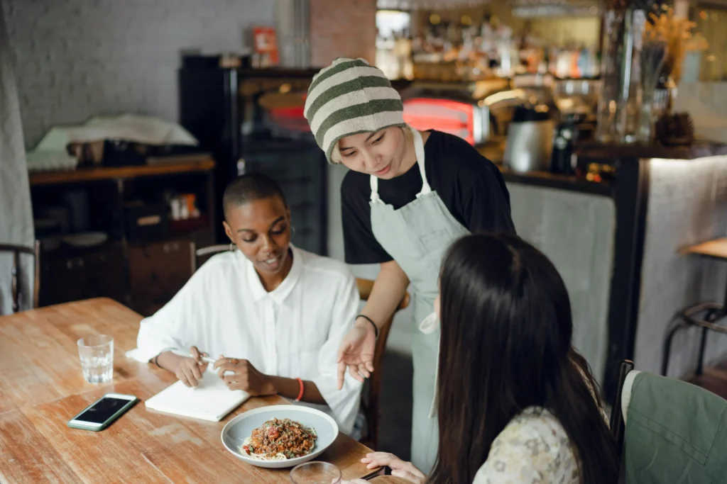 A server in a striped beanie and apron gestures toward a plate of pasta while attending to two customers at a wooden table. The scene captures a focused interaction in a cozy, dimly lit restaurant setting.
