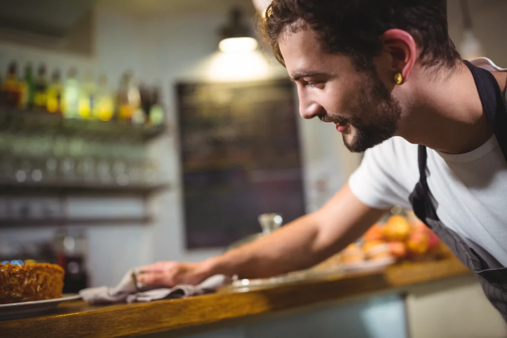 A close-up profile shot shows a smiling male barista with a beard and earring leaning over a wooden cafe counter to wipe it down with a cloth. The background is softly blurred, revealing shelves of bottles, a chalkboard menu, and warm indoor lighting.