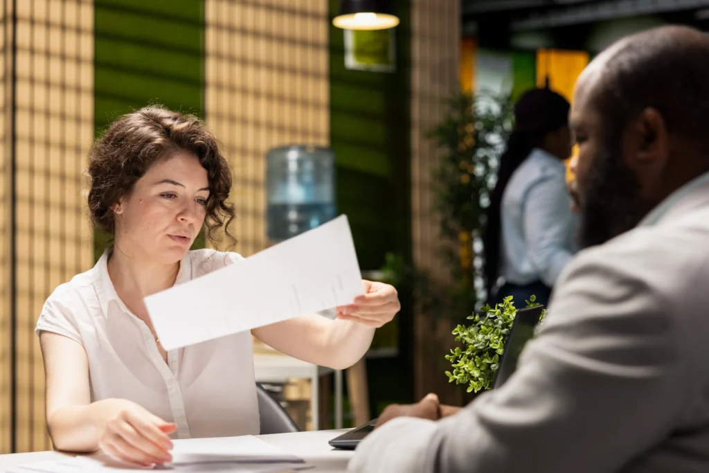 In the image, a woman in a white shirt sits at a desk and hands a document to a man seated opposite her. The setting appears to be a modern office environment with warm lighting and decorative greenery in the background.