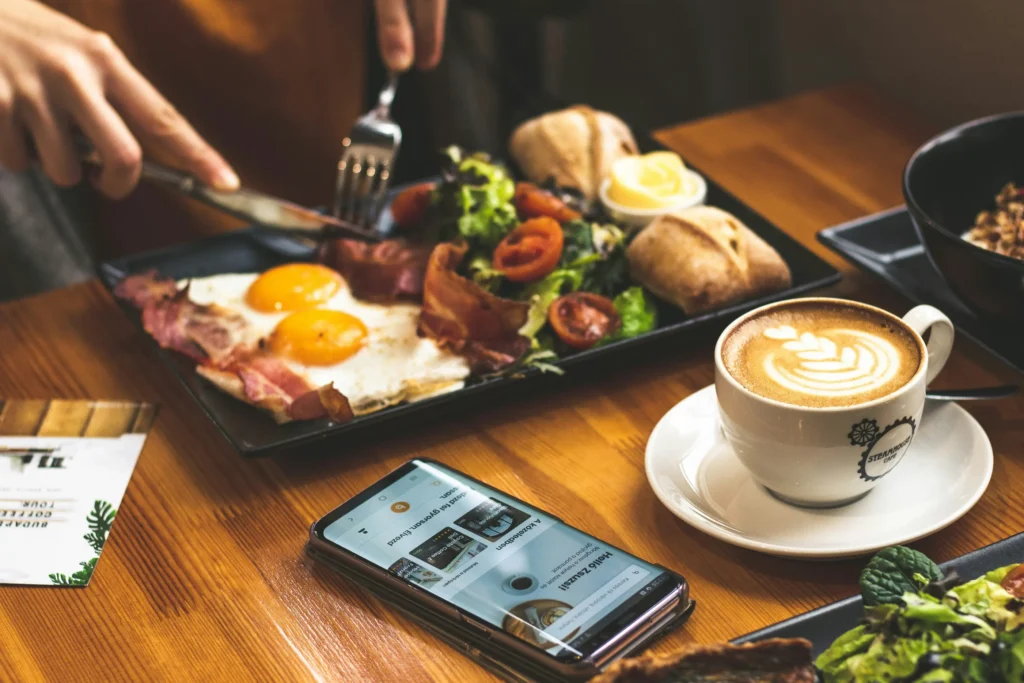 A person uses a knife and fork to eat a breakfast plate featuring sunny-side-up eggs, bacon, a side salad, and bread rolls. A cup of latte art coffee and a smartphone displaying an app sit nearby on the wooden table.