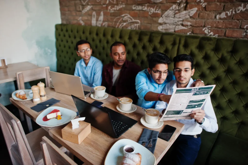 Four South Asian men are gathered around a wooden table in a cafe, working on laptops and looking over a menu together. The table is filled with coffee cups, pastries, a slice of cheesecake, and a smartphone, set against a tufted green booth and a brick wall.