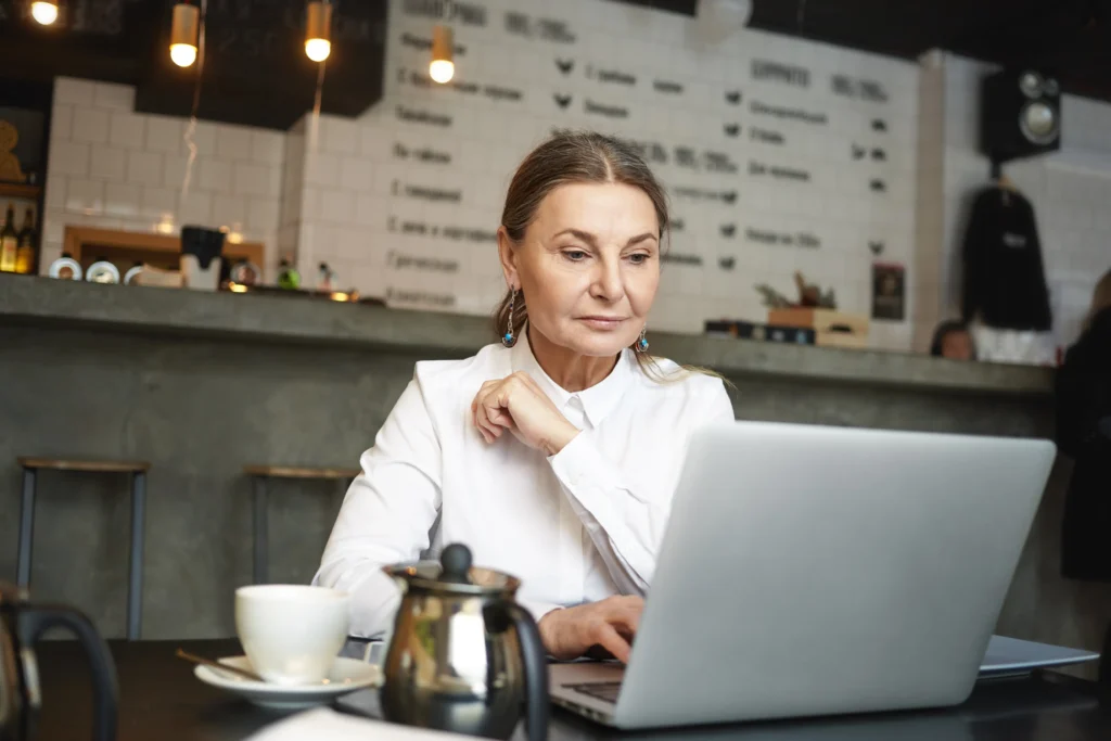 An older woman in a white collared shirt sits at a café table, focused on her laptop with a calm and concentrated expression. In the foreground, a white cup and a metal teapot sit on the table, while a blurred menu board and bar counter create a modern café atmosphere in the background.