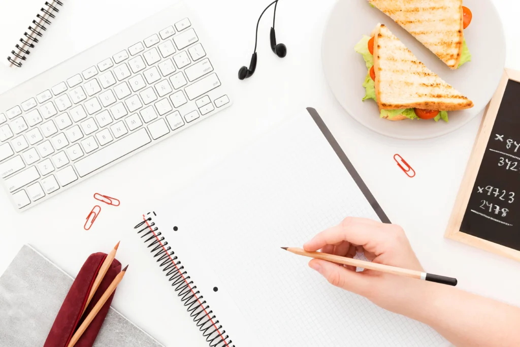 A hand is poised to write in a spiral notebook on a white desk surrounded by a keyboard, earbuds, and a plate of grilled sandwiches. This workspace also features scattered red paperclips, pencils in a pouch, and a small chalkboard displaying math problems.