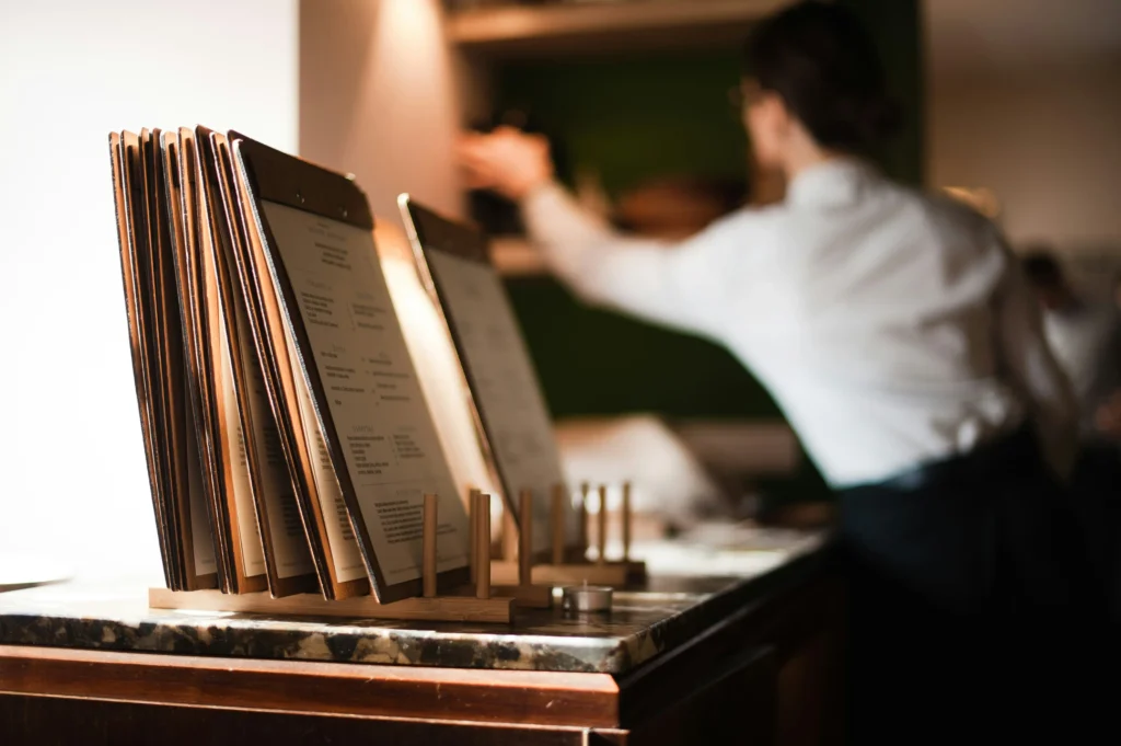 A row of restaurant menus attached to wooden clipboards stands neatly organized in a counter holder, lit by warm ambient light. In the blurred background, a server in a white shirt and black apron reaches toward a shelf.
