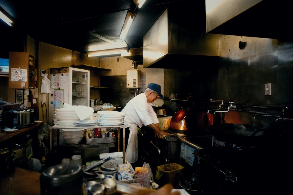 An elderly chef wearing a white uniform and blue cap works in a dimly lit, cluttered commercial kitchen. He stands before a stove where a bright orange flame erupts from a wok, surrounded by stacks of white dishes and industrial kitchen equipment.