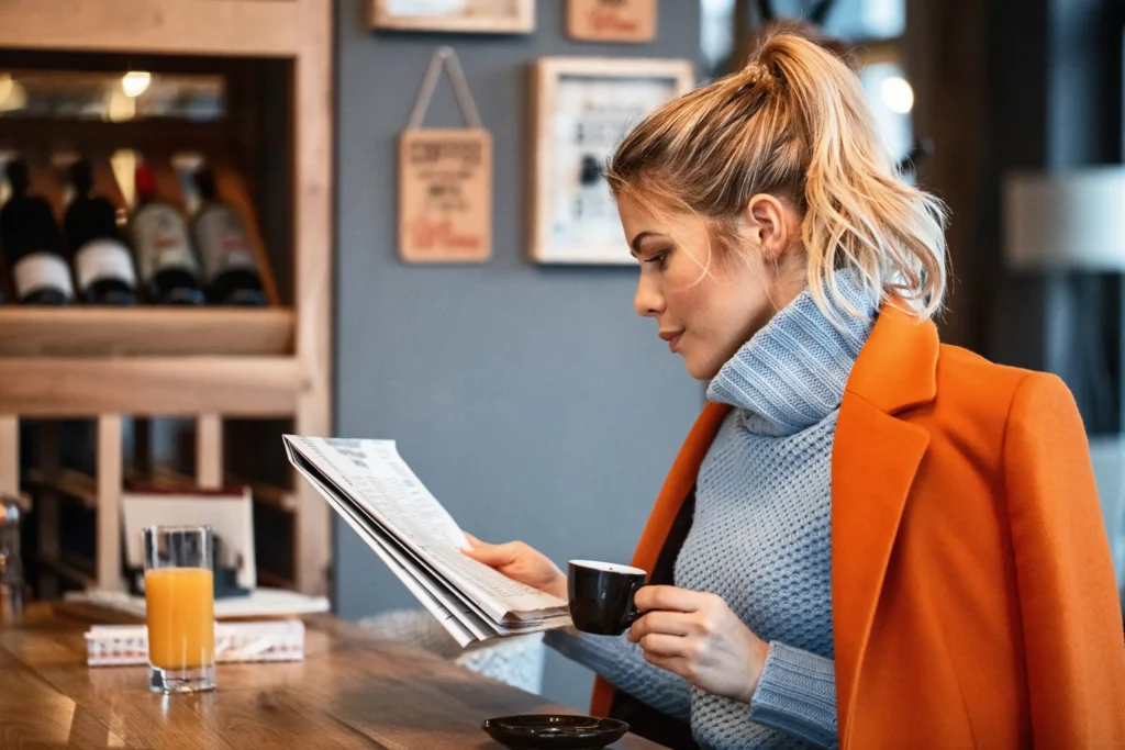 The image depicts a young woman in an orange coat and blue turtleneck reading a newspaper at a cafe table. She sits in profile, holding a small cup of espresso while a glass of orange juice rests nearby on the wooden counter.
